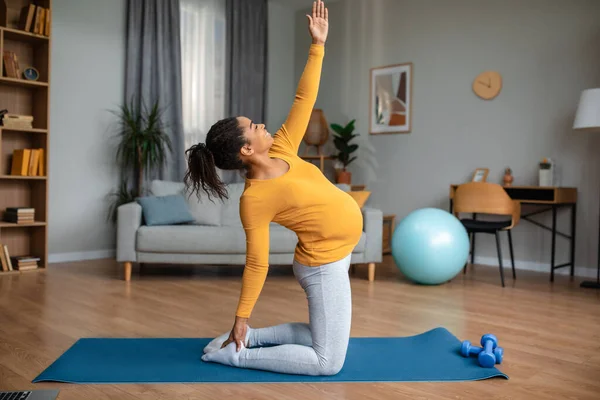Happy smiling young pregnant african american woman practicing yoga on mat, enjoy workout alone in living room interior, profile. Body care during pregnancy, sports at home, expect baby and motherhood