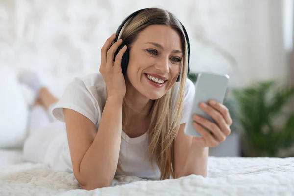 Happy blonde young woman in pajamas laying in bed with smartphone and wireless headphones, watching movie, listening to music or having video chat with friends, weekend at home concept, copy space