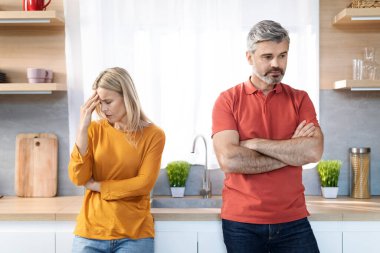 Kızgın orta yaşlı eşler evde kavga ediyor, gri saçlı adam ve sarışın kadın mutfak masasının başında kolları göğsünde duruyor, yana bakıyor, evlilik krizi yaşıyor.
