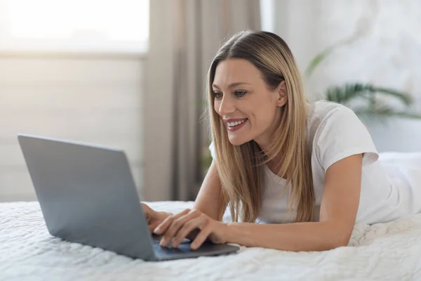 Happy attractive blonde young woman using laptop while staying in bed at weekend at home, typing on computer keyboard and smiling, chatting with guys on dating website, side view, copy space