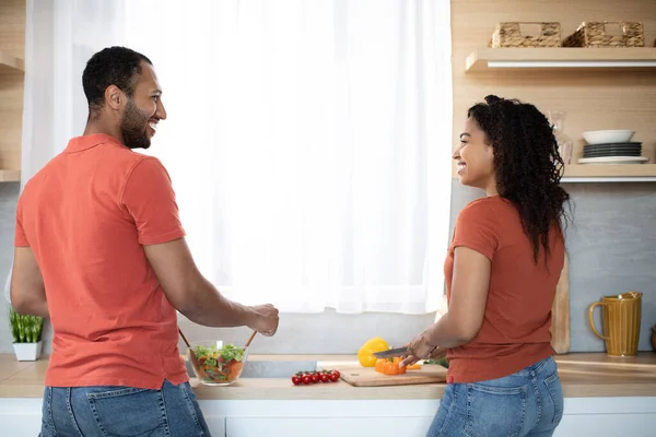 Happy young african american man and woman preparing salad with organic vegetables together at kitchen interior, back. Couple cooking in free time at home, family dinner, love and romance due covid-19