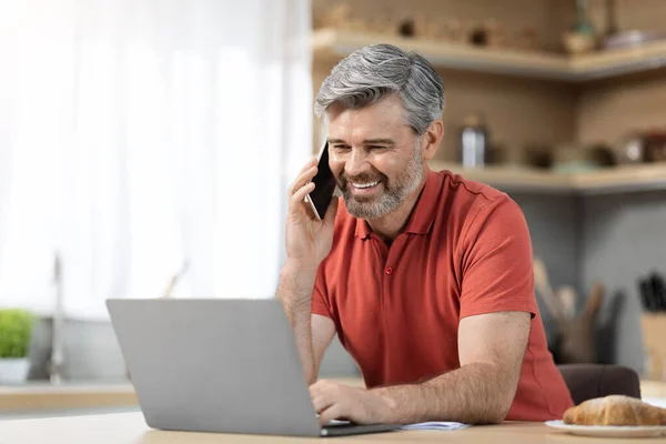 Handsome middle aged businessman working from home, happy grey-haired man having breakfast at kitchen, looking at computer screen and having phone conversation, copy space. Remote job concept