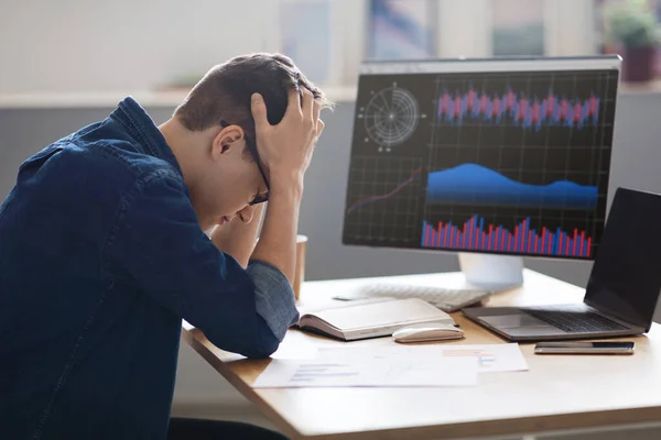 Crypto Crash. Portrait Of Depressed Young Male Employee Sitting At Workplace And Touching Head, Millennial Man Working With Papers And Financial Analytics Charts On Computer Monitor, Side View
