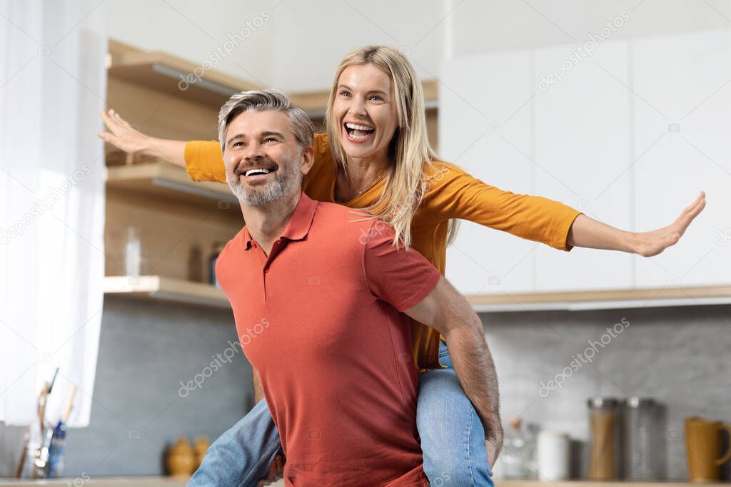 Joyful pretty blonde lady piggybacking her cheerful husband, looking at copy space and smiling, middle aged couple going to cook nice meal together, having fun, kitchen interior