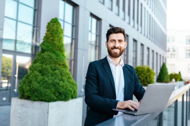 Glad confident european millennial bearded businessman in suit typing on laptop on balcony near modern office building. Work and trade in city. Business remote outdoor, new normal and digital nomad
