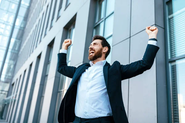 Happy satisfied excited european millennial bearded businessman in suit rejoices in successful deal or win, rises hands up near modern office building, outdoor. Manager, emotions of victory, gestures