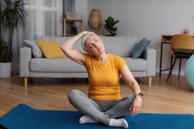 Healthy lifestyle concept. Active senior woman stretching neck, sitting on yoga mat and warming up before domestic workout, training at home in living room
