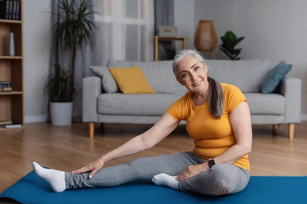 Home workout. Senior woman stretching her leg on yoga mat in living room and smiling at camera, copy space. Positive aged female doing flexibility exercises, keeping fit and healthy