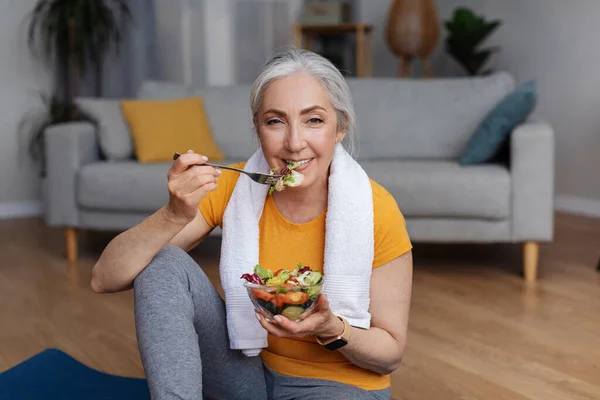Sports and nutrition concept. Happy senior femael leaning eating fresh vegetable salad and looking at camera, sitting on yoga mat at home, enjoying her diet and exercising program