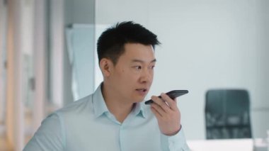 Fast modern communication. Close up portrait of serious mature asian businessman recording voice message on smartphone, standing at office, slow motion
