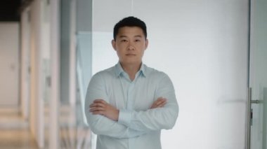 Professionalism concept. Indoors portrait of confident middle aged asian businessman posing with folded arms at office interior, looking at camera, slow motion