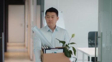 Employment crisis. Upset middle aged asian man manager carrying box with personal belongings and looking sadly at his workplace, beeing fired from job, slow motion, free space