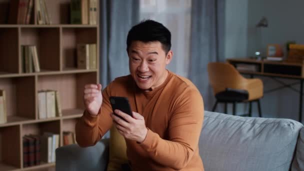 온라인 스마트폰을 보면서 승리를 즐기고 소리지르며 집에서 동작을 동양인 남자의 — 비디오