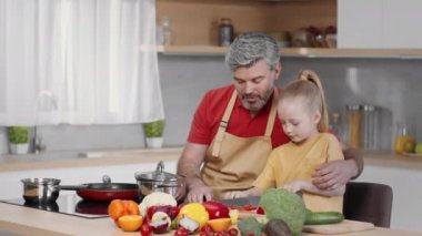 Loving middle aged grey-haired father teaching his little daughter to cut vegetables, preparing dinner together at kitchen, tracking shot, slow motion