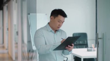 Professional middle aged asian man auditor making inventory at corporate office, making notes in notepad, standing at corridor, slow motion