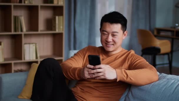 온라인 커뮤니케이션 조심스럽게 이시아인 서핑을 집에서 스마트폰으로 소파에서 휴식을 취하고 — 비디오