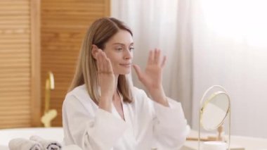 Domestic hairstyling. Pretty positive middle aged lady making ponytail hairdo at home, looking at mirror at bathroom, slow motion, free space