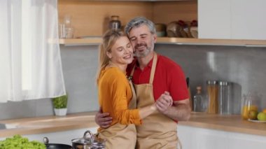 Domestic romance. Happy middle aged man and woman wearing aprons dancing together at kitchen, having fun during cooking dinner, slow motion