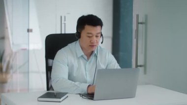 Customer support service. Professional mature asian man operator wearing headset video chatting with client via smartphone, consulting online, sitting at office, slow motion