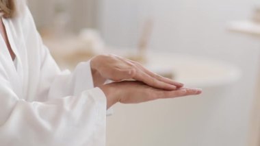 El bakım alışkanlığı. Tanımlanamayan, bornozlu, beyaz bir kadın cildine nemlendirici krem sürüyor, banyoda avuç içini düşünüyor, seçici bir odak noktası var.