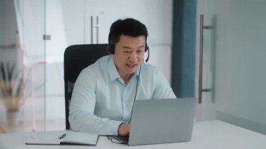 International business concept. Confident middle aged asian businessman wearing headset video conferencing with partners online via laptop, sitting at office, slow motion