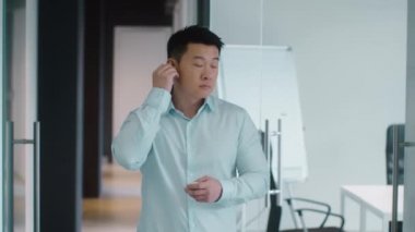 Modern sound technologies. Positive middle aged asian man manager putting earbuds into ears and enjoying great music, standing alone at office corridor, slow motion