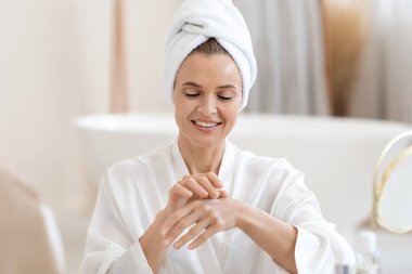 Beyaz bornozlu, saçı türbanlı çekici bir kadın duştan sonra el kremi kullanıyor, ellerine nemlendirici veya besleyici krem sürüyor ve gülümsüyor, fotokopi odası, lüks banyo içi.