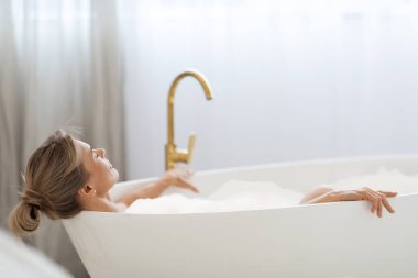 Relaxed blonde lady enjoying hot foamy aromatic bath at home, lying in tub full of water with closed eyes, side view, closeup photo, copy space. Home spa, body care concept