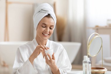 İpek bornoz ve türbanlı güzel bir kadın duştan sonra besleyici yüz kremi kullanıyor, aynaya bakıyor ve gülümsüyor, yüz bakımı rutininden zevk alıyor, kopyalama alanı kullanıyor. Evde yaşlanma karşıtı tedavi