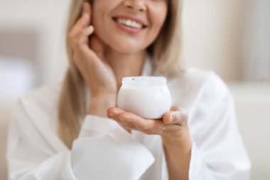 Tanımlanamayan ipek bornozlu, deri nemlendirici kullanan, elinde lüks güzellik ürünü olan beyaz bir kavanoz tutan, gülümseyen, yakın plan çekimli, kırpılmış, kopyalama alanı olan sarışın bir kadın. Yüz bakımı kavramı