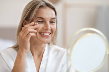 Closeup of cheerful beautiful blonde middle aged lady in silky bathrobe applying moisturizing eye cream, looking at mirror and smiling, enjoying face care routine, copy space