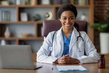 Steteskoplu beyaz cüppeli neşeli doktor Afro-Amerikan kadın klinikte çalışıyor, çalışma masasında oturuyor, dizüstü bilgisayar kullanıyor, hastalarına internetten danışıyor kameraya gülümsüyor, yer kopyalıyor.