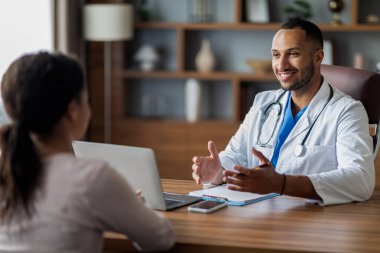 Tanımlanamayan Afro-Amerikan kadın hastalara danışmanlık yapan, doktor tavsiyelerde bulunan, gülümseyen ve el kol hareketi yapan, klinikteki dizüstü bilgisayarla çalışma masasında oturan yakışıklı Arap doktor.