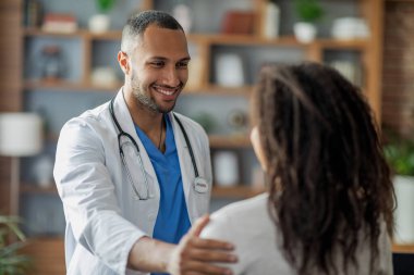 Sağlık sigortası konsepti. Çekici, çekici, ortadoğulu genç doktor. Kadın hastasını selamlıyor. Afrikalı Amerikalı kadın, omzuna dokunup gülümsüyor.
