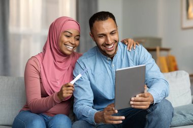 Mutlu siyahi Müslüman çift, evde koltukta otururken hamilelik testi ve dijital tablet yaptırıyor, gülümseyen İslami eşler çocuk doğurmaya hazırlanıyor, internetten sağlık sigortası talep ediyor,