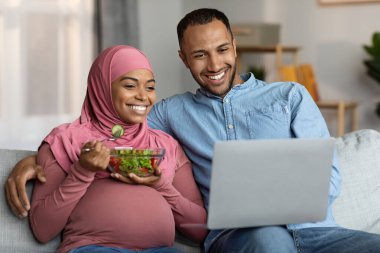 Mutlu Müslüman çift oturma odasında dizüstü bilgisayarla rahatlıyor, gülümseyen İslami eşler evde bebek istirahati bekliyor, Kanepede film izliyor, tesettürlü kadın sebze yiyor,