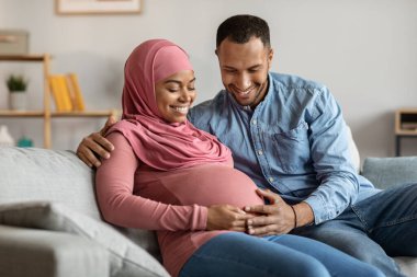 Mutlu siyah islami bir çiftin portresi bebek beklerken koltukta dinleniyor, gülümseyen hamile bir çift evde kaynaşıyor, sevgi dolu koca karına dokunuyor, yakın plan
