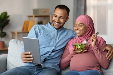 Hamile Çiftler İçin Boş Zaman. Siyahi İslami Eşler Koltukta Dijital Tabletle Rahatlıyor, Müslüman Çiftler Film İzliyor veya Modern Gadget 'ta Alışveriş Yapıyor, Türbanlı Kadın Bekliyor Salata Yiyor