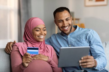 Mutlu siyahi çift dijital tablet ve kredi kartıyla internetten satın alırken evde koltukta dinlenirken gülümseyen İslami eşlerin internet alışverişinden keyif almasını bekliyor.