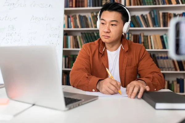 Japanese Male Taking Notes Looking At Laptop Computer Learning Or Teaching Online Wearing Wireless Headphones Sitting In Modern Classroom. Internet Education Concept