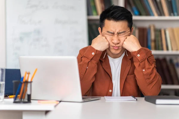 Asian Male Teacher Sitting With Eyes Closed Near Laptop Tired Of Computer Work Having Professional Burnout In Modern Classroom. Online Education Issues Concept