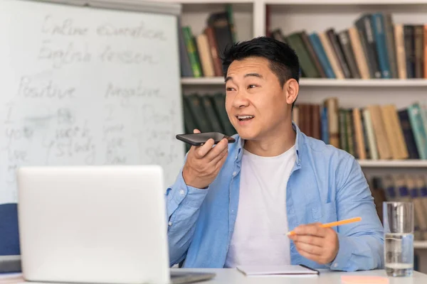 Asian Male Tutor Using Voice Search Application On Phone Sitting Near Laptop At Workplace. Teacher Having Online Class Remotely. Modern Education And Technology Concept
