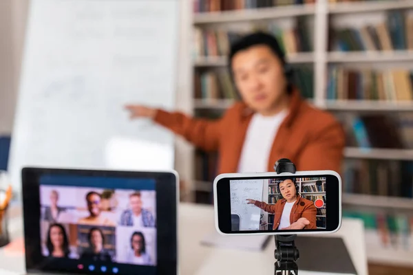 E-Teaching. Japanese Teacher Man Filming Online Lecture On Cellphone And Laptop Computer Pointing At Whiteboard Making Video Call Sitting In Classroom. Remote Education. Shallow Depth