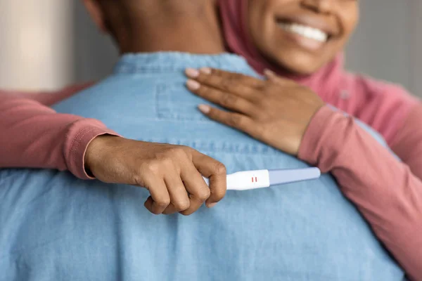 Expecting a baby. Happy black islamic woman hugging husband and holding positive pregnancy test, joyful pregnant muslim female embracing spouse, celebrating becoming parents, cropped shot, closeup