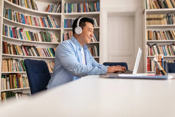 Korean Man Wearing Headphones Using Laptop Typing Sitting In Library Indoors. Businessman Working Online Via Computer. Technology And Business Concept. Side View Shot