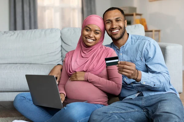 Online Payments. Pregnant Black Islamic Spouses With Laptop And Credit Card Posing In Living Room Interior, Smiling Muslim Couple Enjoying Shopping In Internet While Awaiting Baby, Copy Space