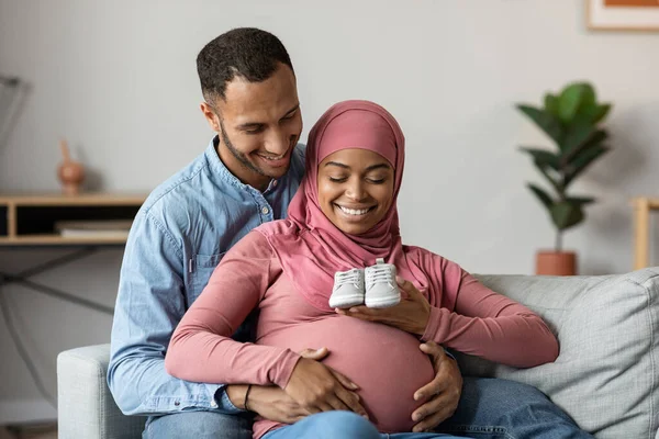 Sonriente Pareja Negra Islámica Embarazada Sentada Sofá Sosteniendo Zapatos Bebé — Foto de Stock