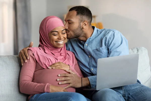 Romantik İslamcı eşler evde dizüstü bilgisayarla bebek bekliyor, siyah kocasını seven hamile karısını öpüyor, tesettürde yan yana yatarken hafifçe karnına dokunuyor.