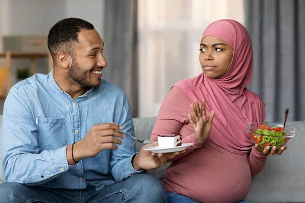 Black man offering cake to his pregnant muslim wife while she eating salad, serious islamic lady in hijab refusing sweets, choosing vegetables over dessert, having healthy nutrition during pregnancy