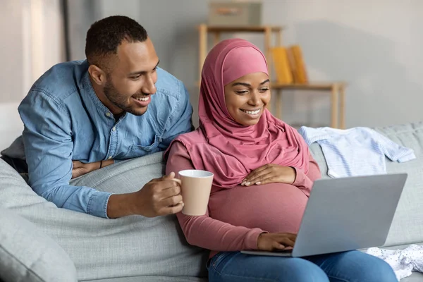 Hamile siyahi kadın ve kocası evde dizüstü bilgisayar kullanırken, mutlu İslami eşler oturma odasında dinlenirken internetten alışveriş yaparken, ev içi tatilin keyfini çıkarırken,